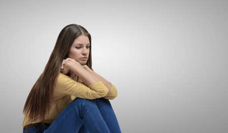A young beautiful woman in casual clothing sitting placed her elbows on her knees on a white backgroundの写真素材