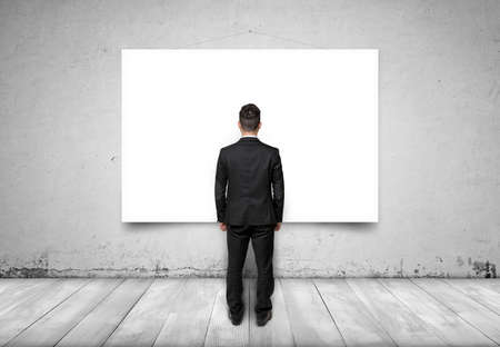 Back view of a businessman standing on a wooden floor looking at a white empty canvas on a concrete wallの写真素材