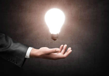 A hand of a businessman in a gray suit holding a glowing light bulb in his hand on concrete wall