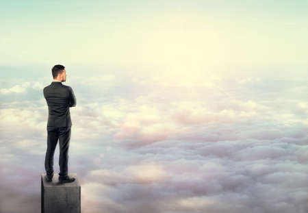 Young man in business suit, standing on a concrete column above the clouds.の写真素材