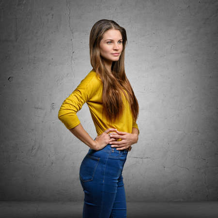 Charming young woman wearing yellow blouse and jeans, standing sideways with hands on waistの写真素材