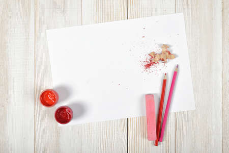 Top view of designer workplace equipped with red and pink gouache jars, colored pencils, chalk, shavings and white paper on light wooden surface.の写真素材
