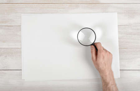 Hand of man holding the magnifying glass over white paper lying on wooden surface. Top view.の写真素材