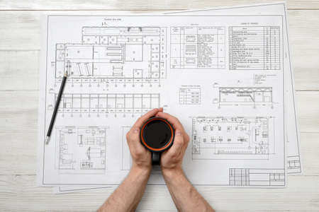 Close-up hands of man holding a cup of coffee over drawing layout in top view. Workplace of architect or constructor. Engineering work. Architect drawing. Uplifting mood. Coffee break. Conducive working environment. Increasing productivity in the morningsの写真素材
