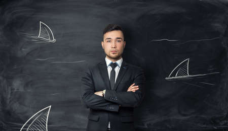 Businessman stands on background with sketches of shark fins. Symbol of fearlessness, sophisticated cunning, power and danger. Dangerous world of business and finance. Surrounded by enemies.の写真素材