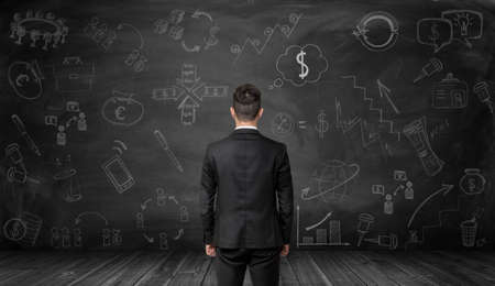 Back view of businessman looking at a black wall with hand-drawn graphics pictures and diagrams related to economics and statistics. Making money. Increase in profits. Prosperous business. Srategic calculation and research. Financial and business concept.の写真素材