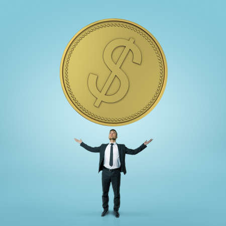Businessman stands rising his hands towards big coin isolated on blue background.の写真素材