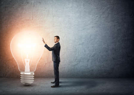 Businessman stands touching big glowing light bulb. Ideas nad concepts. Creativity and inspiration. Signs and symbols. Intellectual job. Business innovations.の写真素材