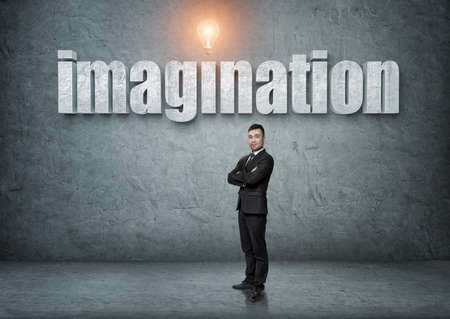 Businessman stands with his arms folded with big 3d 'imagination' word above. Ideas and concepts. Business staff. Office clothes. Intelligence and creativeness.の写真素材