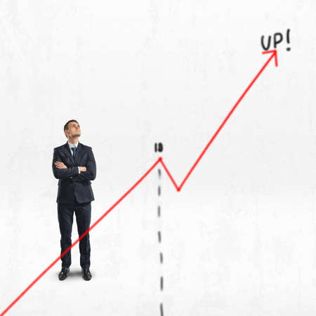 Businessman stands with folded arms looking upwards behind big rising diagram isolated on white background.の写真素材