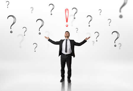 Businessman stands with raised hands looking upwards at an exclamation mark surrounded by question marks isolated on white background.の写真素材