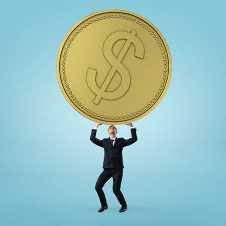 Businessman stands and holding heavy, big golden coin on blue background.の写真素材