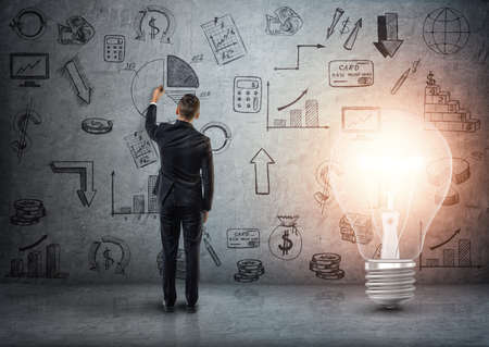 Back view of a businessman drawing business doodles on concrete wall with big glowing light bulb beside him.の写真素材