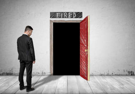 Back view of a businessman standing in front of an open door with darkness behind it and word 'fired' above it.の写真素材
