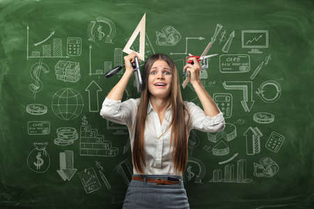 Cropped portrait of young businesswoman in stress holding office supplies over her head on the background of a chalkboard with business doodles. Problems at work. All hand's job.の写真素材