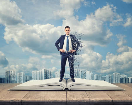 Front view of a businessman standing on an open book with letters covering his right side on the background of urban scene and cloudy blue sky.の写真素材