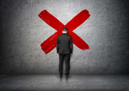 Back view of a businessman lean with his forehead to concrete wall with big red cross in the middle. Problems at work. Lack of inspiration. Difficulties and troubles.の写真素材