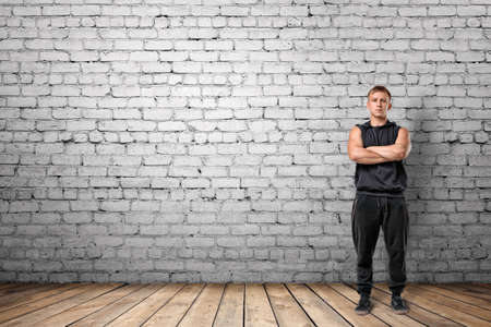 Front view of muscled young man standing with his arms folded on the background of white brick wall. Wellbeing. Muscleman. Workout and fitness.の写真素材