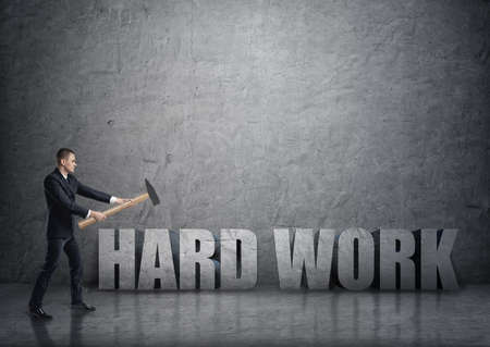 Side view of young businessman crashing 3D concrete 'hard work' words with a hammer. Efficiency at work. Succesful management. Business staff.の写真素材