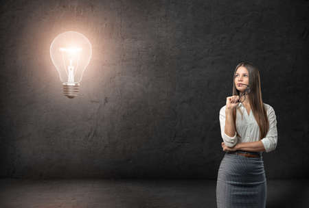 Young businesswoman with glasses looking thoughtfully on the glowing light bulb. New ideas. Inspiration.の写真素材
