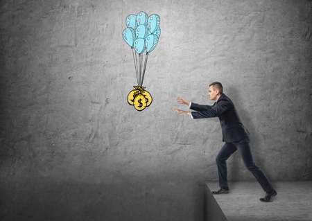 Businessman standing on the edge reaching for sacks of money hanged on balloons. Lust of money....