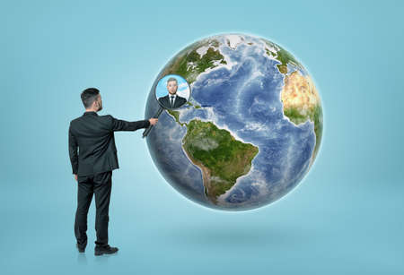 Back view of a businessman looking through magnifying glass on the Earth and seeing a portrait of a man. Global communication. International business.の写真素材