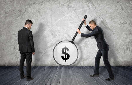Two businessmen: the right one enlarging dollar sigh with big magnifier, the left one looking at it. Profit and success. Teamwork. Experience and knowledge.の写真素材