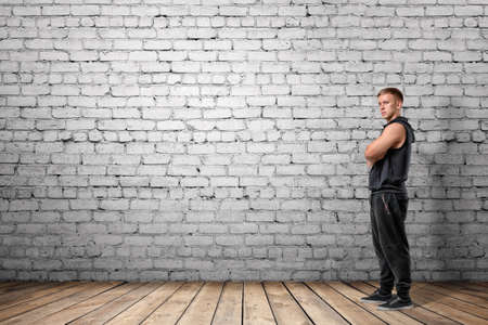 Front view of muscled young man standing with his arms folded looking at the camera, on the background of white brick wall. Wellbeing. Muscleman. Workout and fitness.の写真素材