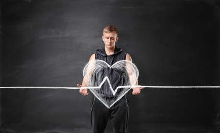 Muscled young man holding big drawn beating heart on the background of a blackboard. Healthcare and...