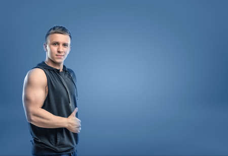 Croppeed portrait of muscled young man showing his bicep and thumbs up on blue background. Self-improvement. Athlete and bodybuilder. Workout and fitness.の写真素材