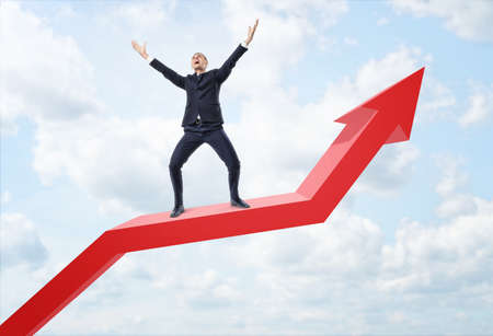 Businessman expressing happy and standing on a big red line graph with an upturned arrow on a background of the sky. Achieving success. Personal development and career progress. Growth of income.の写真素材