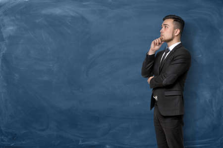 A businessman in a black suit standing with his hand on his forehead on the dark blue background. Business planning. Looking for opportunities. Management challenges.の写真素材
