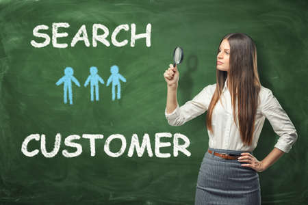 A beautiful businesswoman standing and looking through the magnifying glass near the green wall with the words 'search customer' and tiny blue humans drawn on it. Account manager. Business expansion. Search of opportunities. CRM.の写真素材