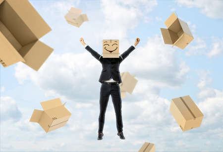 A businessman wears a box on his head with a funny face painted on it, lifts his hands up like being happy and empty boxes flying away from him on a sky background. Business success. Face masks. Hidden emotions.の写真素材