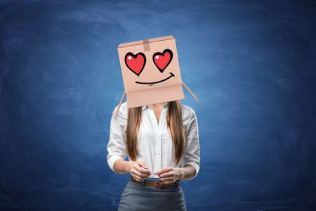 Young beautiful businesswoman with cardboard box on her head with drawn smiley face with hearts instead of eyes. Anonymous person. Falling in love. Office romance. Love customers.の写真素材