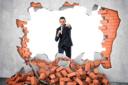A businessman standing near the hole in the wall which he has just made with a hard punch, a pile of red bricks are lying beneath. Overcoming obstacles. Business issues. Economic development.の写真素材