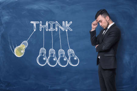 Businessman standing with hand on his forehead near word 'think' drawn on blue chalkboard. Business...