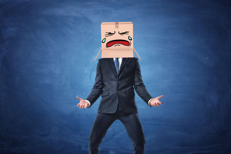 Businessman wearing cardboard box with drawn crying face on his head. on blue chalkboard background. Anonymous person. Issues of small business. Bear losses.の写真素材