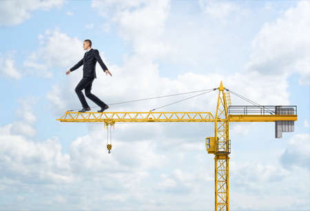 A businessman walking on the yellow construction crane, trying to balance himself and get to the end of the jib, on the sky background. Business difficulties and risks. Overcoming obstacles. Construction equipment and machinery. House-building and reconstの写真素材