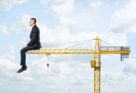 A huge businessman sitting on the end of a crane on the sky background. Dreaming about career growth. Building industry. Supervising manpower.の写真素材