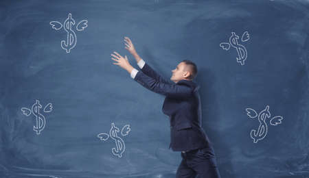 A businessman trying to catch the dollar signs with wings drawn on the blue blackboard. Making money. Success and profit. Business conceptの写真素材