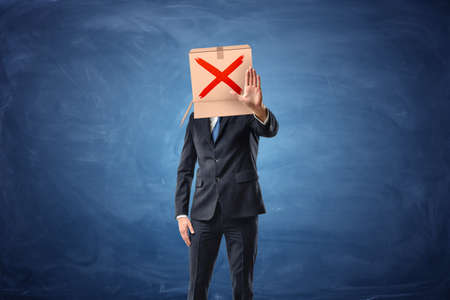 Businessman is wearing cardboard box with drawn red cross sign on it and making stop sign by his hand, on blue chalkboard background. Don't do it. Restrictions and limitations. Serious risk.の写真素材