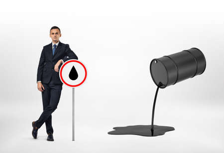 A businessman leaning on the road sign with the oil drop depicted on it, and oil pouring out of the black barrel, isolated on the white background. Oil and petroleum products. Black gold. Resource extraction.の写真素材