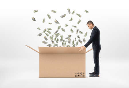 A businessman in a suit standing near the open big beige cardboard mail box with dollar bills falling into it, on the white background. Make profits. Business development. Rise in profitability.の写真素材