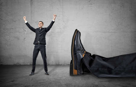 A small businessman standing with his arms up and smiling happily near a giant leg in trousers and shoe lying near him, on the concrete gray background. Winning contest. Best in field. David and Goliath. Small company wins big one.の写真素材