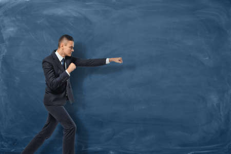 A businessman in a black suit standing as if he is going to punch something invisible near the blue chalkboard. Aggressive marketing strategies. Business development. Successful leader.の写真素材