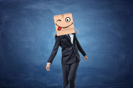 Businessman wearing cardboard box with drawn winking face on his head on blue chalkboard background. Time to have fun. Crazy idea. Attracting clients.の写真素材