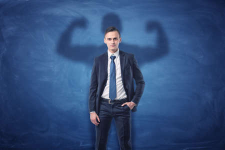Businessman is casting shadow of big strong muscular man showing his biceps. on blue chalkboard background. Inner strength. Leadership qualities. Business development.の写真素材