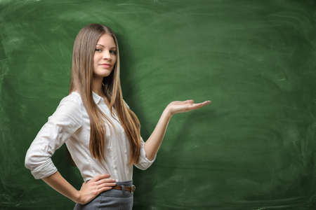 A beautiful young businesswoman holding her open palm and showing at the empty area on the green chalkboard behind her. Concept of advertisement product. Business development. Advertising department.の写真素材