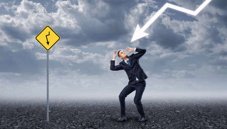Businessman standing on a rocky ground frightened by a lightning, near yellow road sign with the same arrow painted on it. Business crisis. Temporary difficulties. Emotional pressure.の写真素材